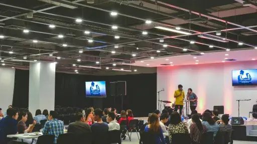 A large indoor event with attendees seated at tables facing a stage where two individuals are presenting.