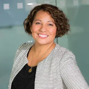 A person in professional attire standing in front of a glass wall with frosted square patterns, wearing a striped blazer and smiling.