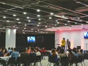 A large indoor event with attendees seated at tables facing a stage where two individuals are presenting.
