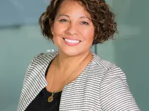 A person in professional attire standing in front of a glass wall with frosted square patterns, wearing a striped blazer and smiling.