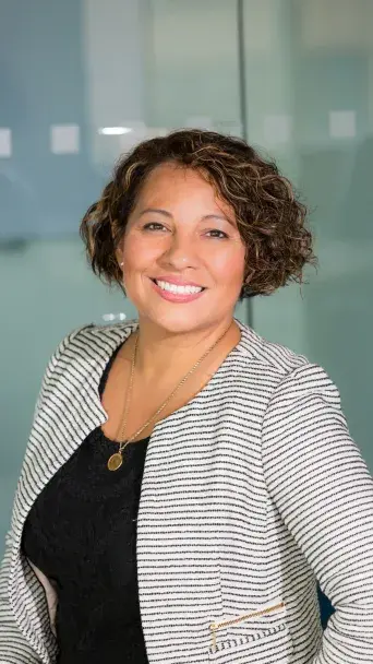 A person in professional attire standing in front of a glass wall with frosted square patterns, wearing a striped blazer and smiling.